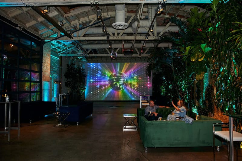 Warehouse party space with a rainbow disco ball video wall, green sofas and tall plants