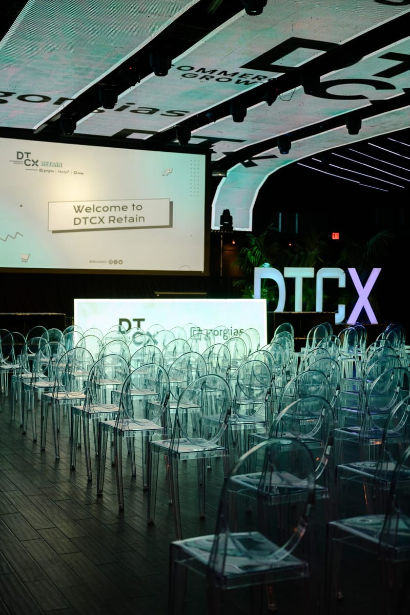 Rows of clear chairs facing a Welcome to DTCX Retain screen under curved LED panels