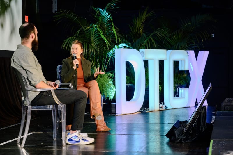 Two speakers in clear chairs holding a fireside chat beside giant light-up DTCX letters and palms