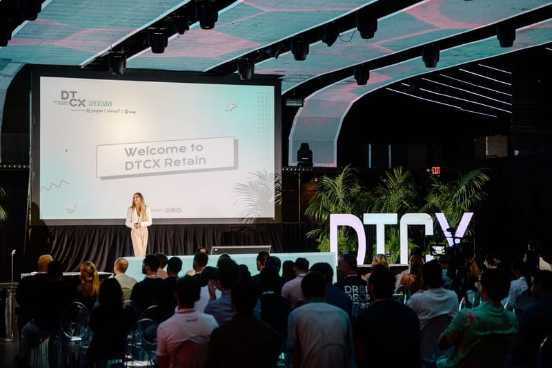 Speaker in white on stage below a Welcome to DTCX Retain screen, facing a seated audience
