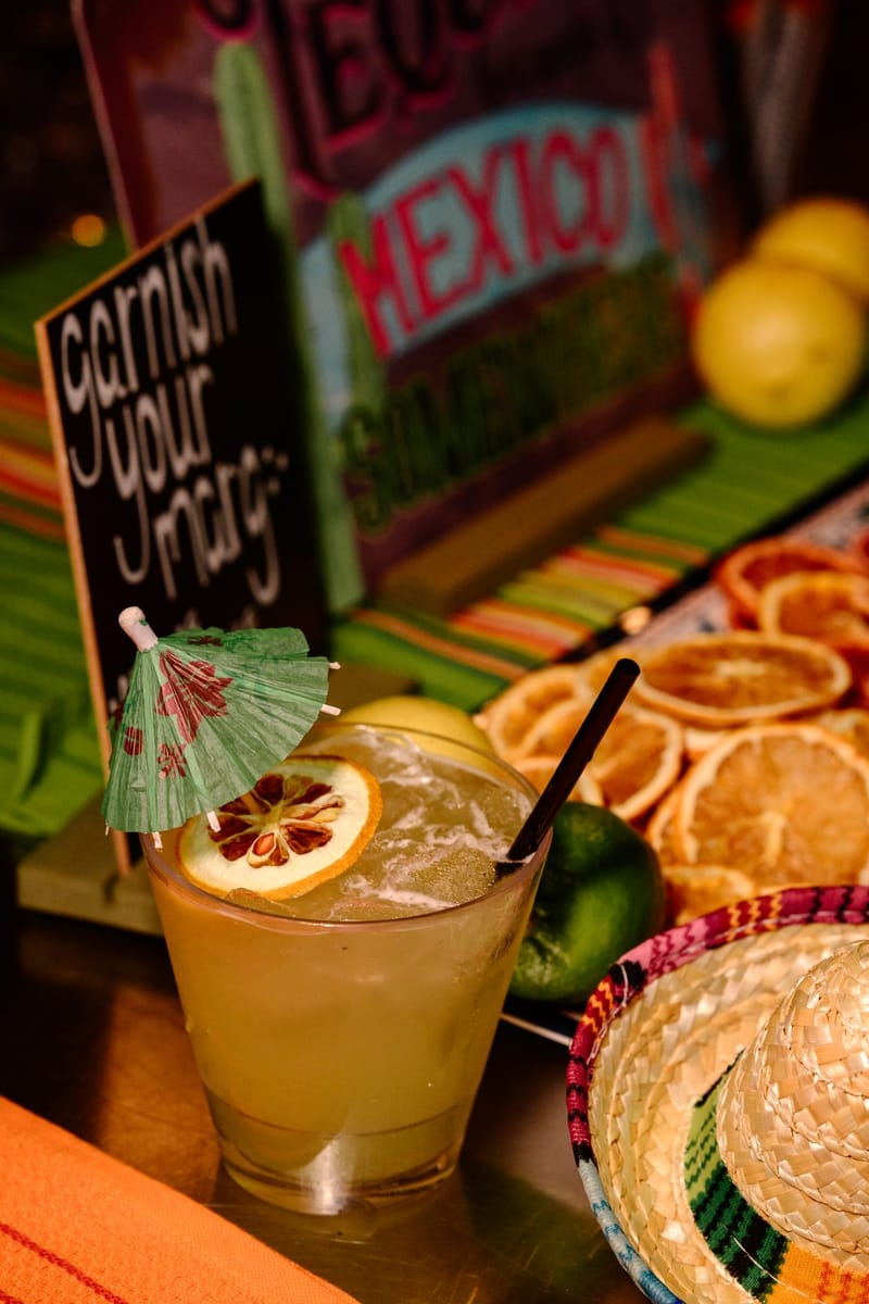 Margarita with a paper umbrella and dried citrus beside a garnish your marg sign