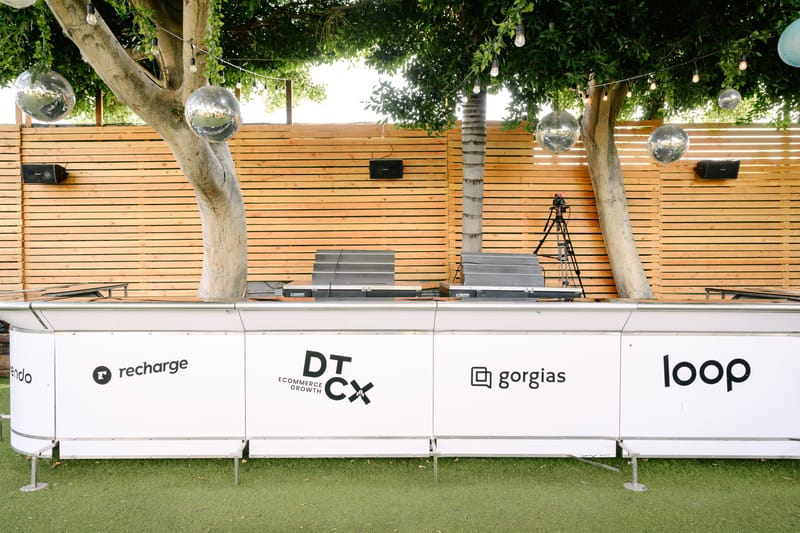 White outdoor bar printed with recharge, DTCX, gorgias and loop logos under trees hung with disco balls
