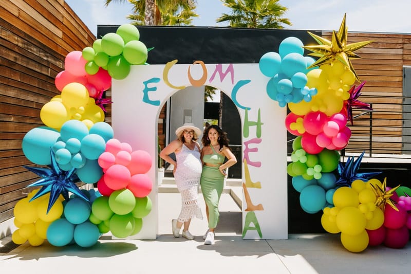 Two women posing in the white arched ECOMCHELLA entrance framed by bright balloon garlands and gold star balloons