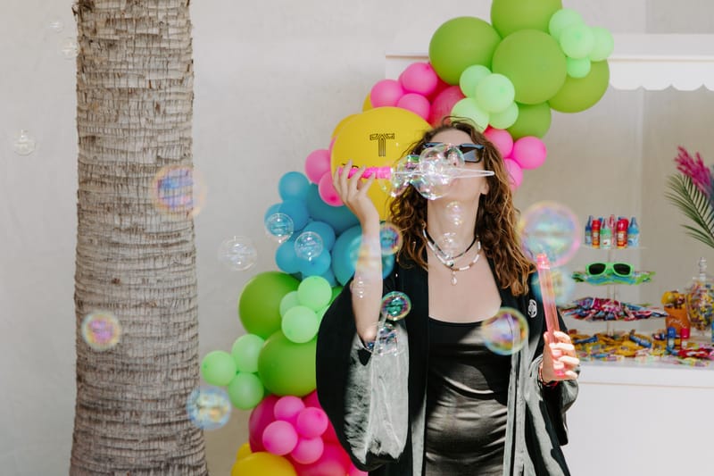 Woman blowing soap bubbles beside a green and pink balloon garland near a candy table