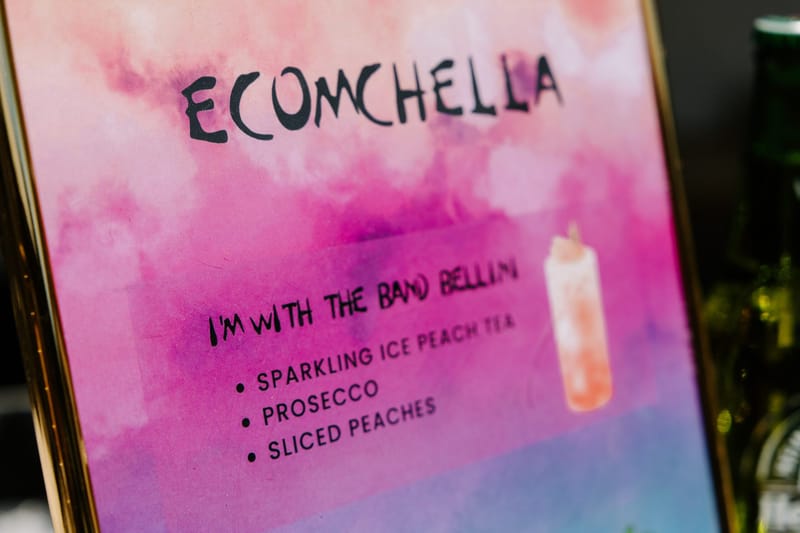 Watercolor ECOMCHELLA sign listing the I'm With the Band Bellini with sparkling ice peach tea, prosecco and sliced peaches
