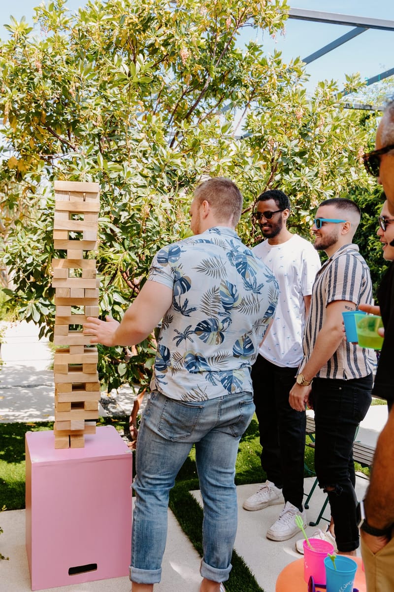 Guest pulling a block from a giant wooden block tower on a pink stand while two friends watch