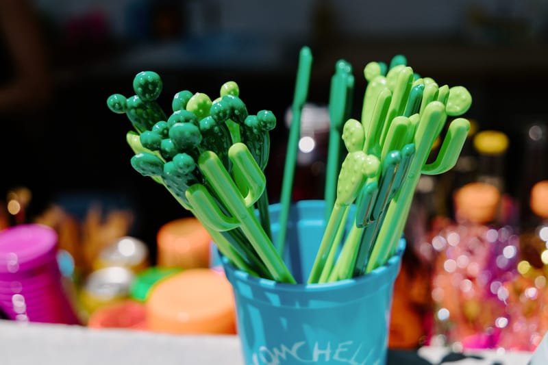 Cactus-shaped drink picks in a teal ECOMCHELLA cup with colorful bottles blurred behind