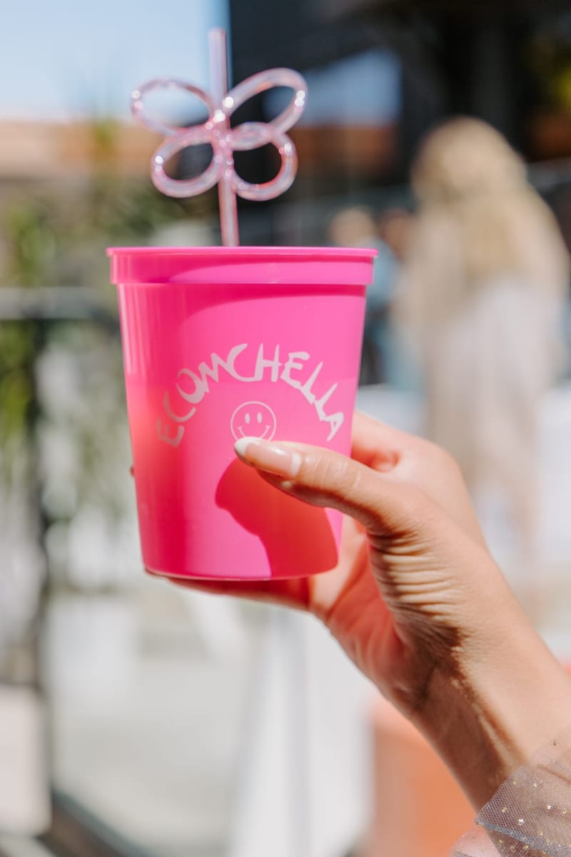 Hand holding up a pink ECOMCHELLA cup with a smiley face and a pink butterfly straw