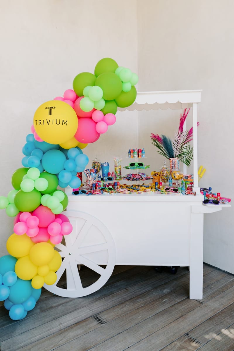White candy cart on wheels beside a TRIVIUM balloon in a colorful balloon garland