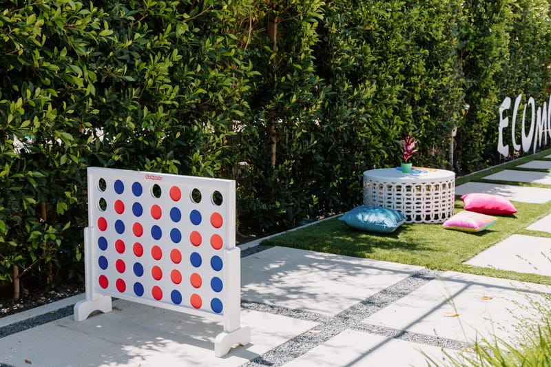 Giant red and blue four-in-a-row game on a lawn, with ECOMCHELLA letters in the hedge behind
