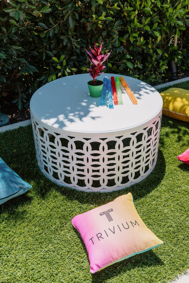 Pink TRIVIUM pillow on artificial grass beside a white lattice side table with a small plant