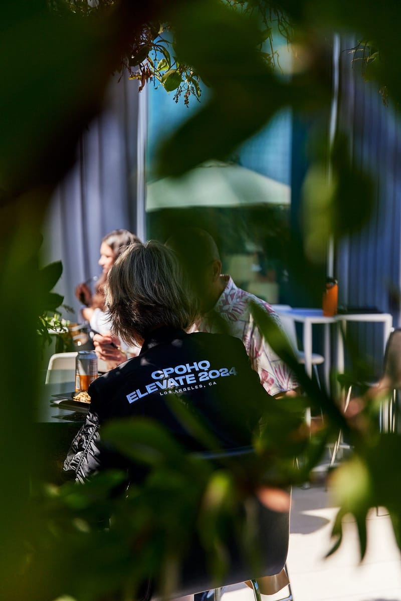 A guest in a CPOHQ Elevate 2024 Los Angeles jacket seated outdoors, seen through leaves