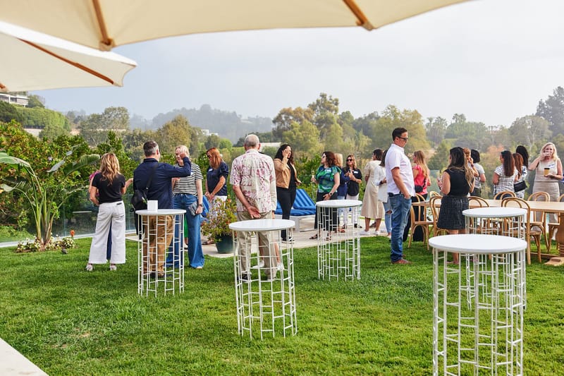 Guests standing at white high-top tables on a lawn with a hillside view, under an umbrella