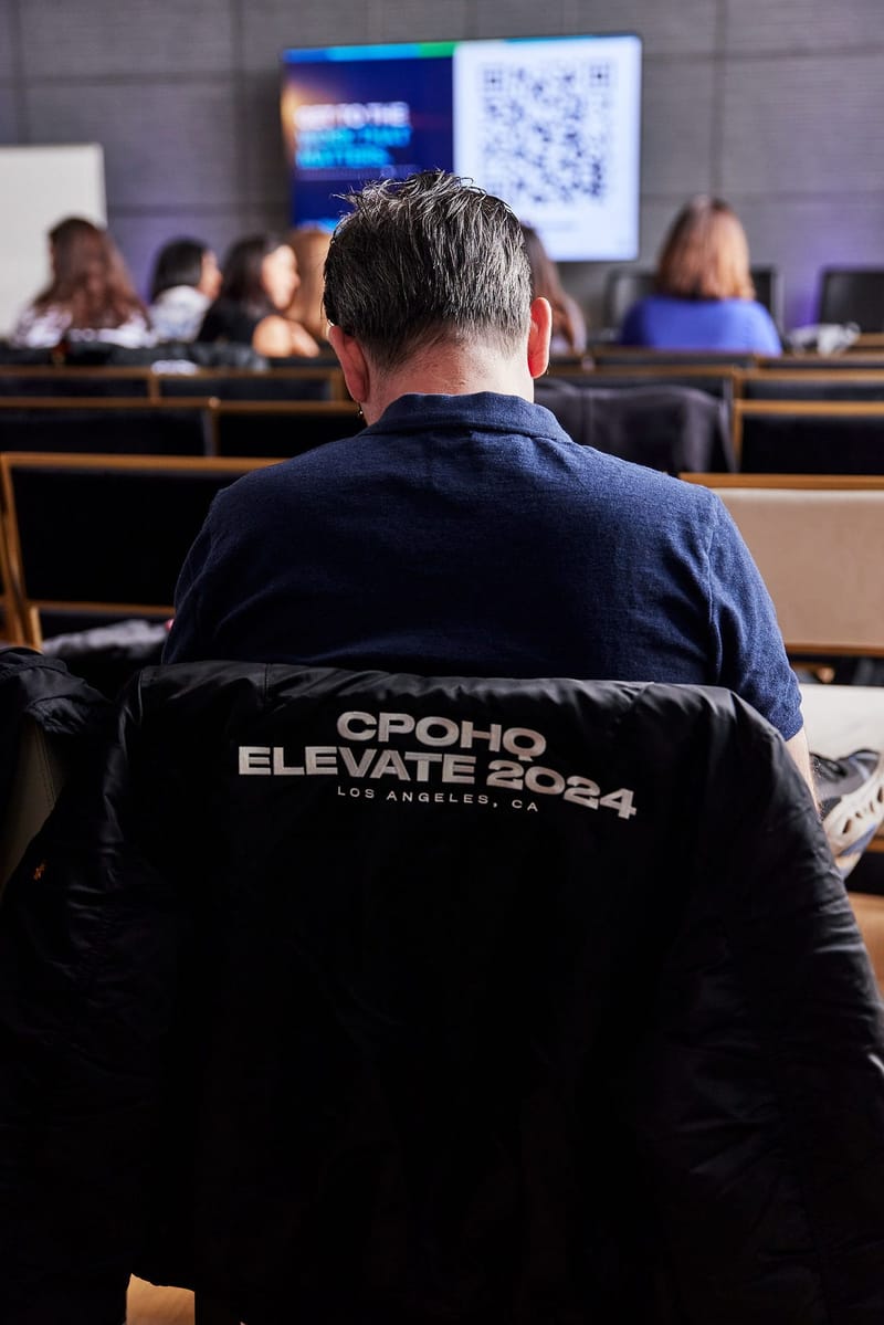 Seated audience facing a screen with a QR code, a CPOHQ Elevate 2024 jacket over a chair