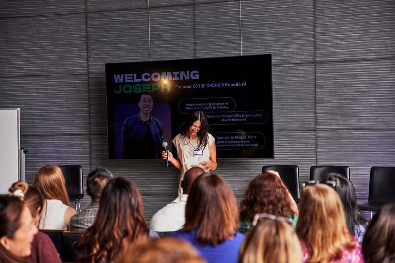 A speaker with a microphone and notes in front of a welcome slide, facing a seated audience
