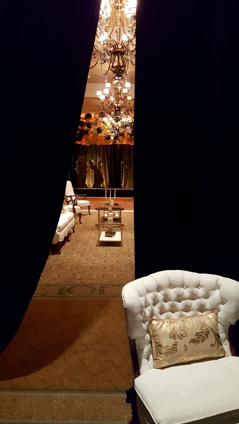 View between black curtains into a lounge with a chandelier and a white tufted chair with a gold pillow