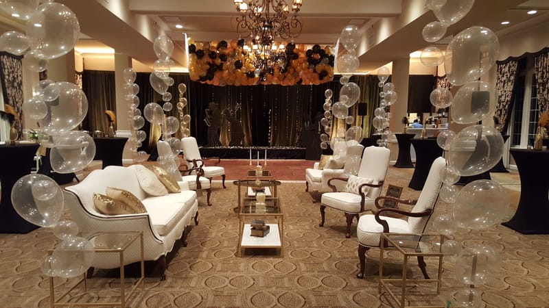 Lounge of white sofas and tufted armchairs around low tables under a chandelier, with clear balloon columns