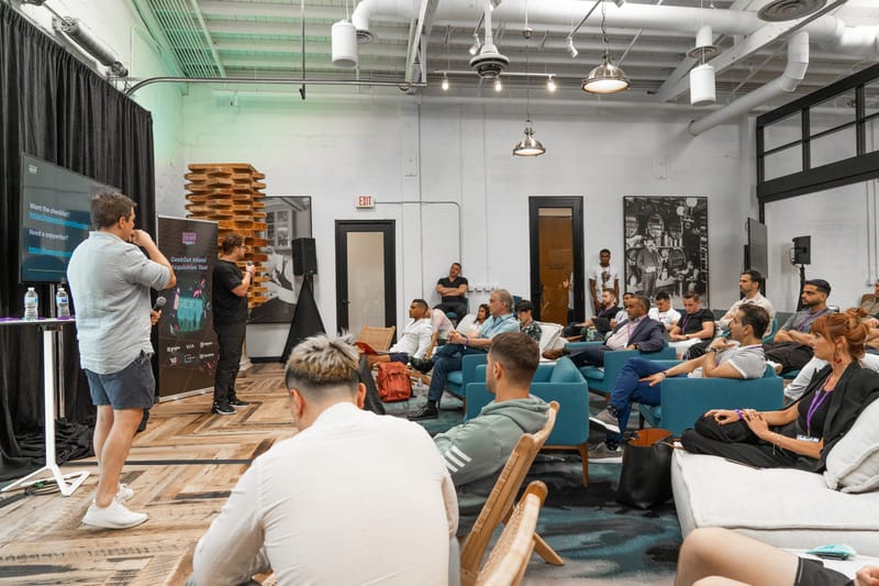 Speaker addressing guests in teal armchairs and white sofas in a white room with a GeekOut banner