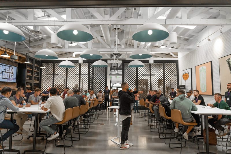 Speaker standing among attendees at white tables under teal pendant lamps in a bright hall