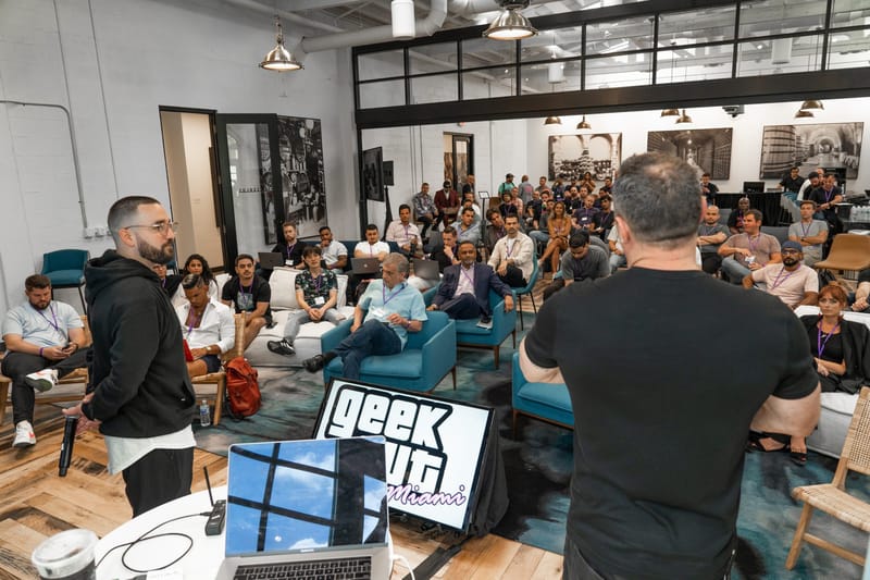Two speakers facing a full room of seated attendees, with a small Geek Out Miami screen in the foreground