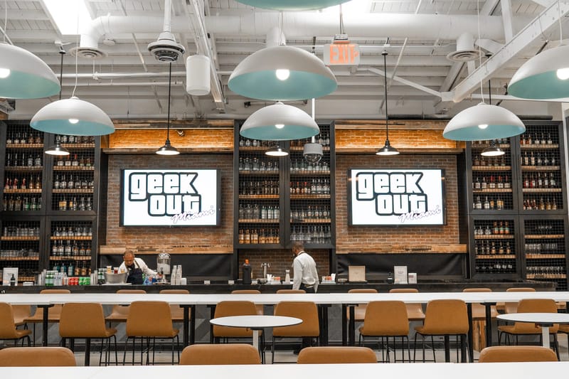 Bar with shelves of bottles and two screens reading geek out Miami above a row of white tables