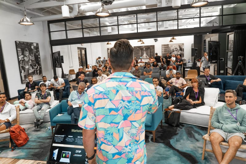 Speaker in a patterned shirt seen from behind, facing a full lounge of attendees in teal armchairs