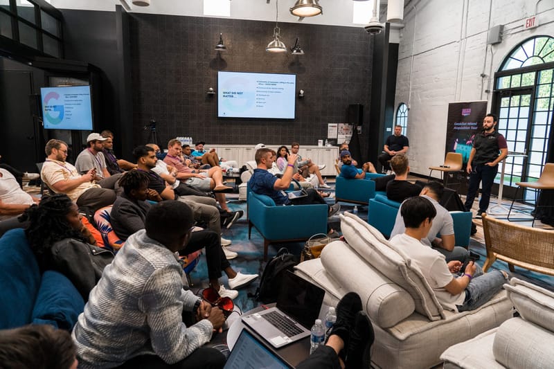 Attendees on sofas and armchairs with laptops watching presentation screens on a dark tiled wall
