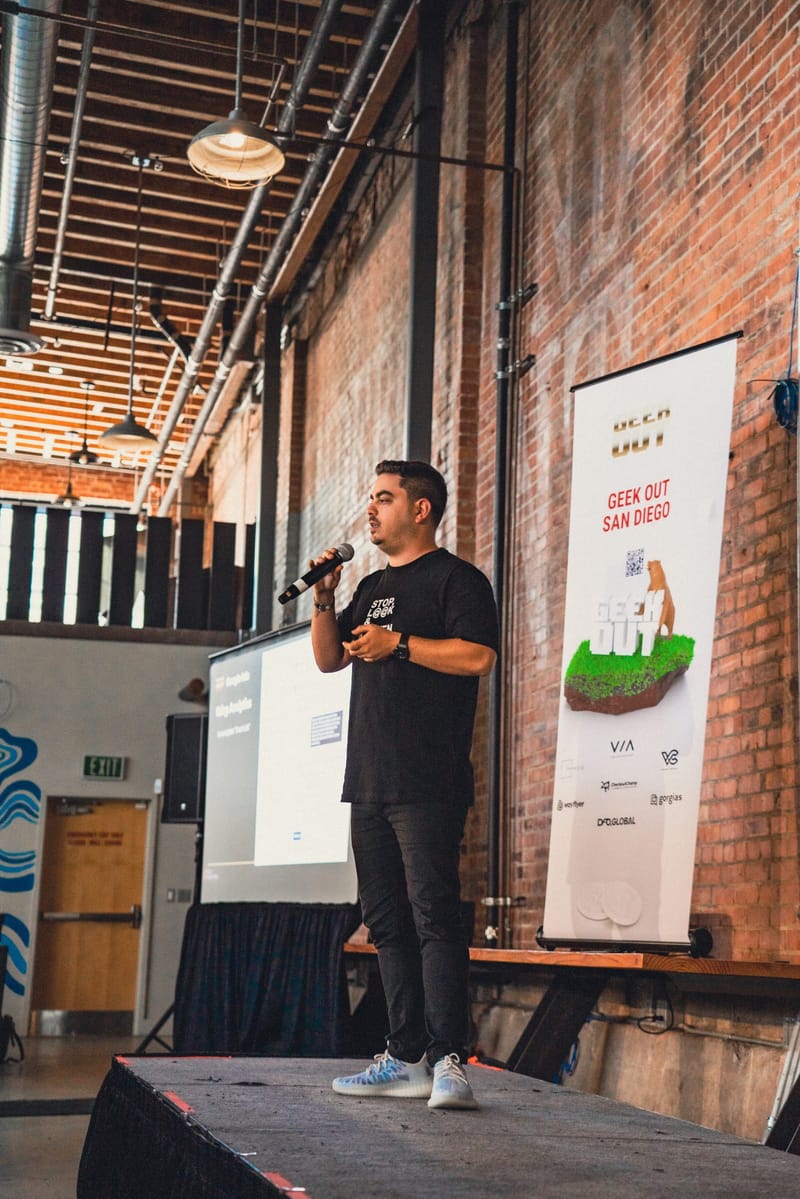 Speaker with a microphone on a low stage against brick, beside a Geek Out San Diego banner with sponsor logos
