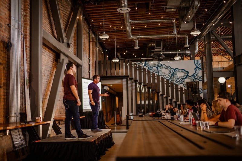 Two speakers on a low stage beside long wooden tables of attendees in a hall with steel beams