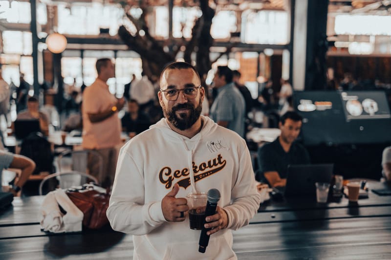 Man in a white Geekout San Diego hoodie giving a thumbs up while holding a microphone and a drink