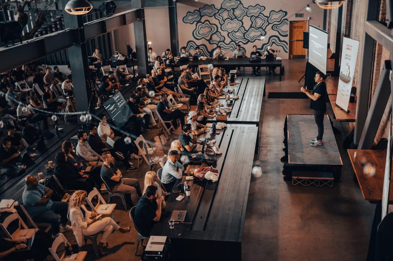 Wide overhead view of a packed hall of attendees at long tables and folding chairs facing a low stage