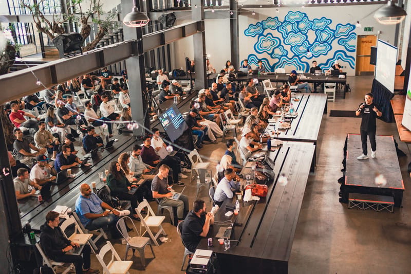 Speaker with a microphone addressing attendees with laptops at long tables, with a blue swirl mural on the wall