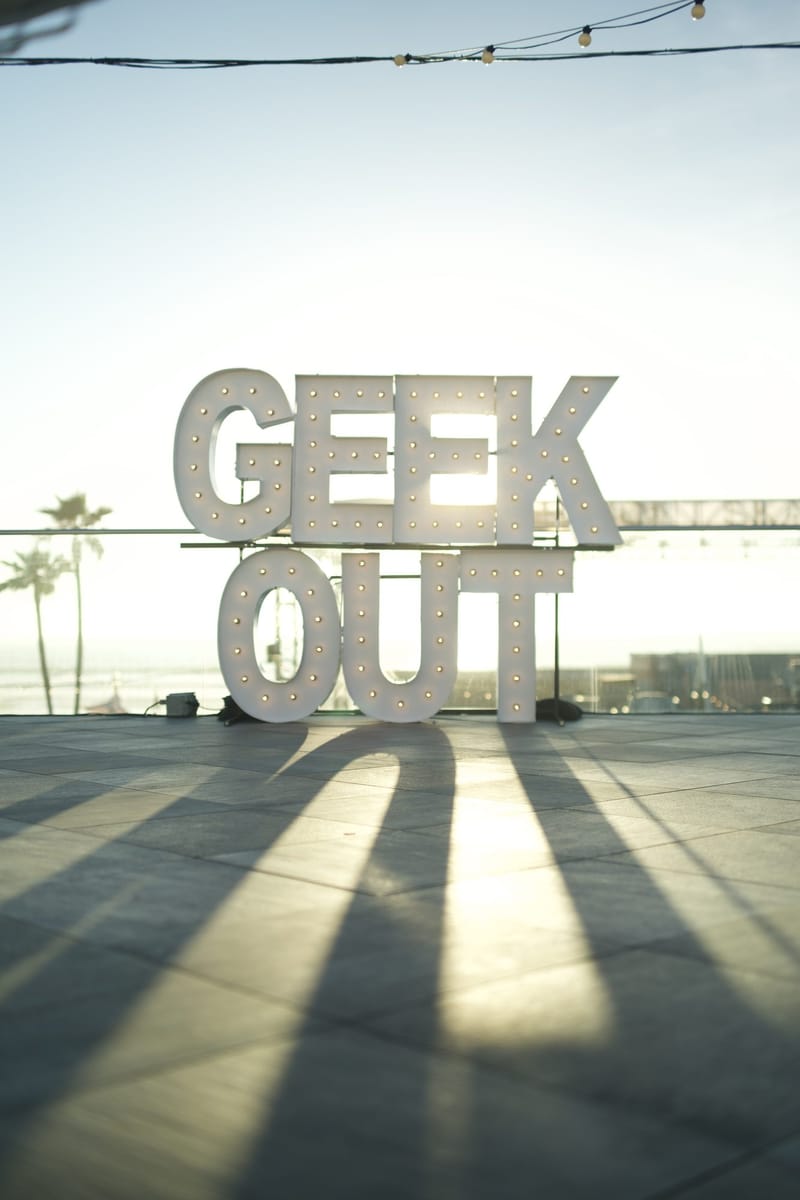 Marquee letters spelling GEEK OUT on an empty terrace, backlit by the sun, with palm trees behind