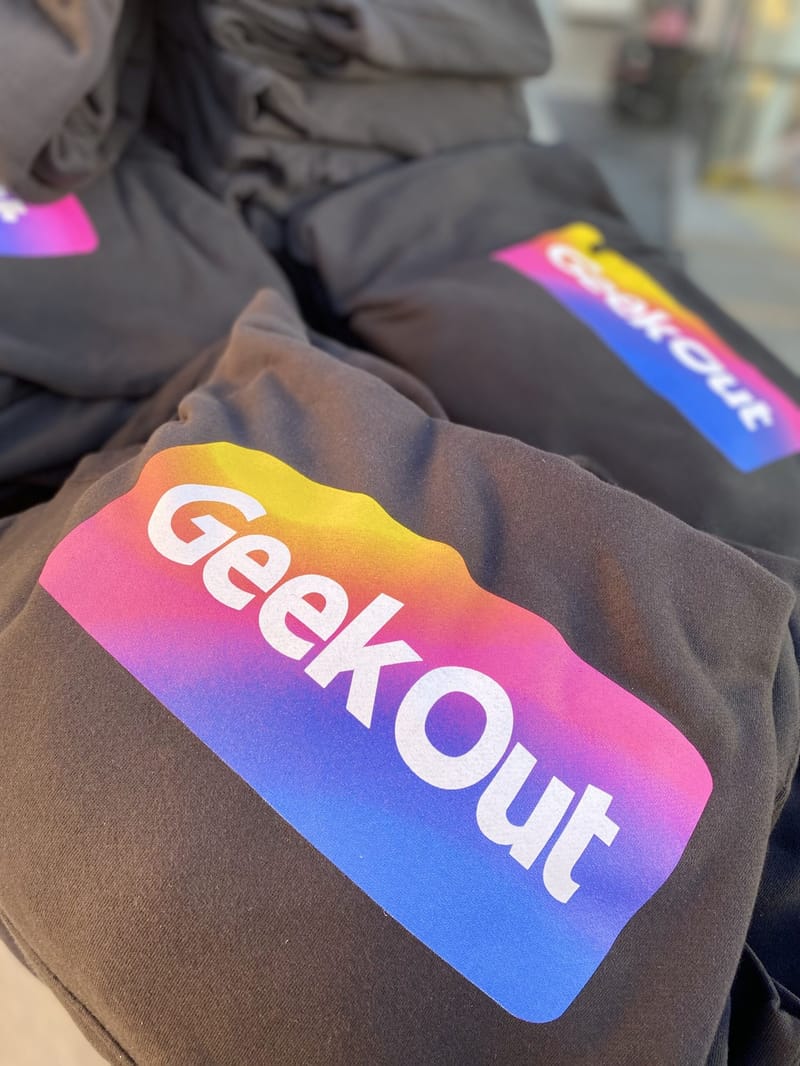 Folded black sweatshirts printed with a pink, yellow and blue GeekOut logo