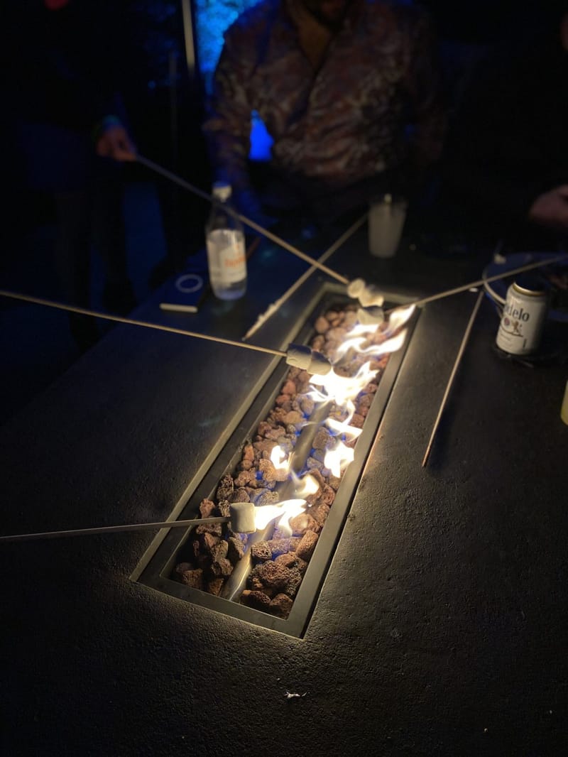 Guests holding marshmallows on long sticks over a fire table filled with lava rocks