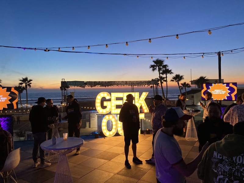 Guests in silhouette at dusk around lit GEEK OUT letters under string lights, with the ocean behind