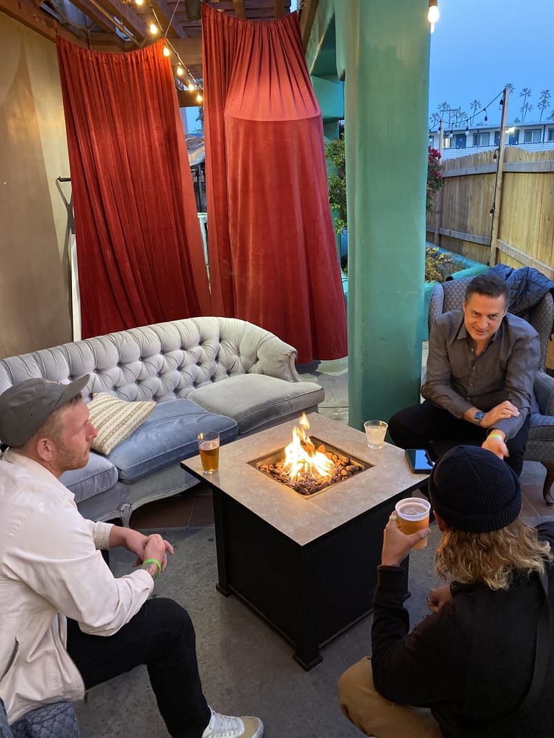 Four guests with drinks around a square fire pit beside a tufted sofa and red velvet curtains