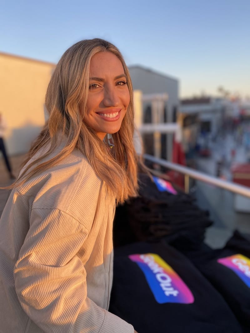 A smiling woman in a cream jacket beside a table of black GeekOut sweatshirts on a sunny terrace