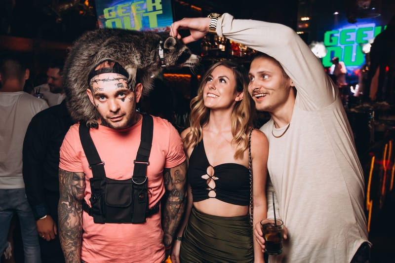 Three guests smiling up at a raccoon perched on one man's head, with GEEK OUT screens behind