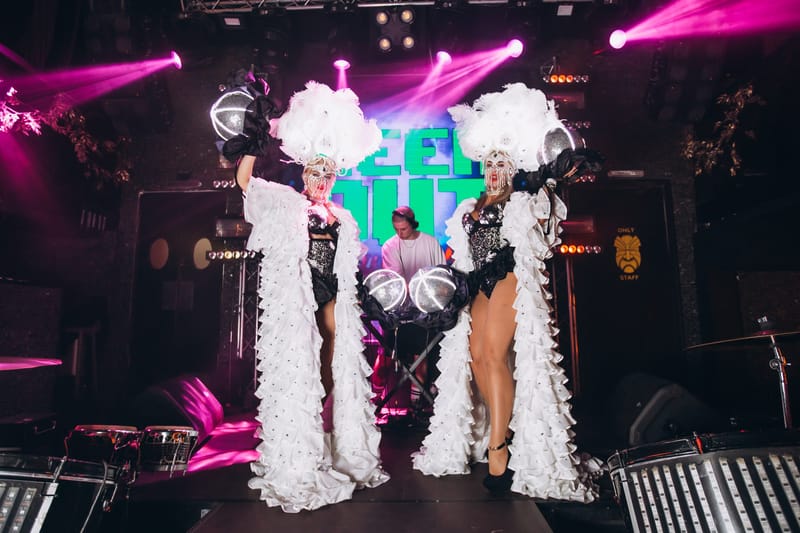 Two performers in white feathered costumes posing on stage under pink spotlights