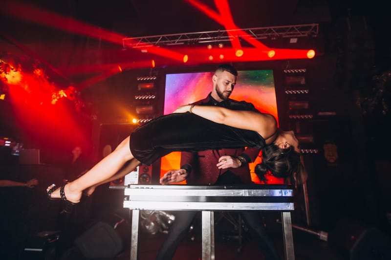 Performer holding a woman in a black dress horizontally above a metal table under red lights