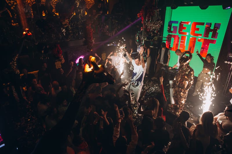 Performer in a mirrored suit on a stage with sparklers beside a green GEEK OUT screen above the crowd