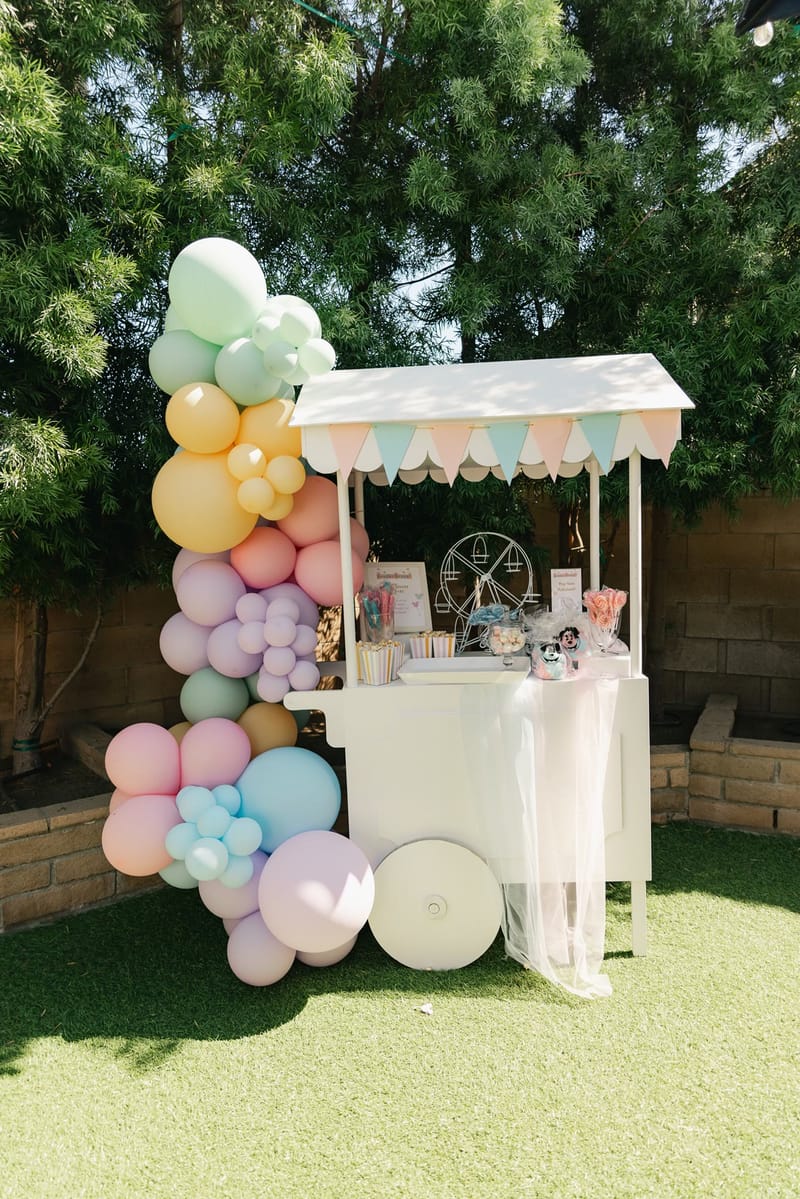 White candy cart with a scalloped pastel awning beside a pastel balloon garland on the lawn