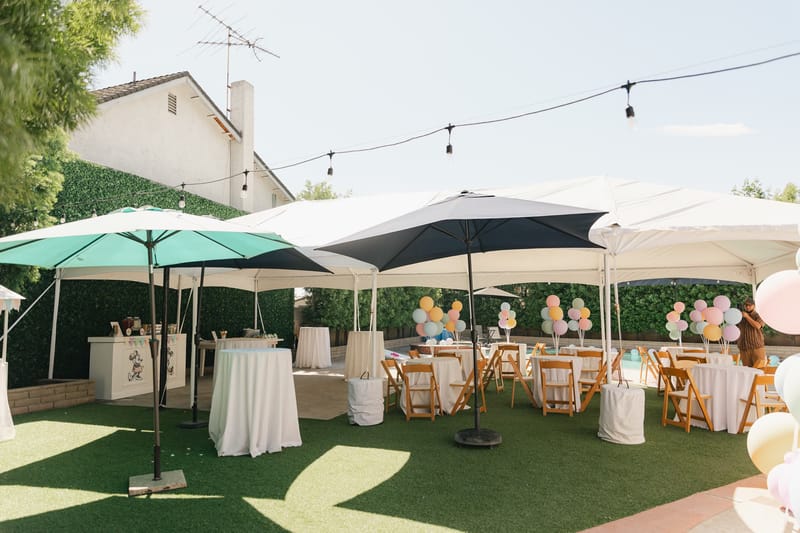White tents and teal umbrellas over round tables on turf in the backyard