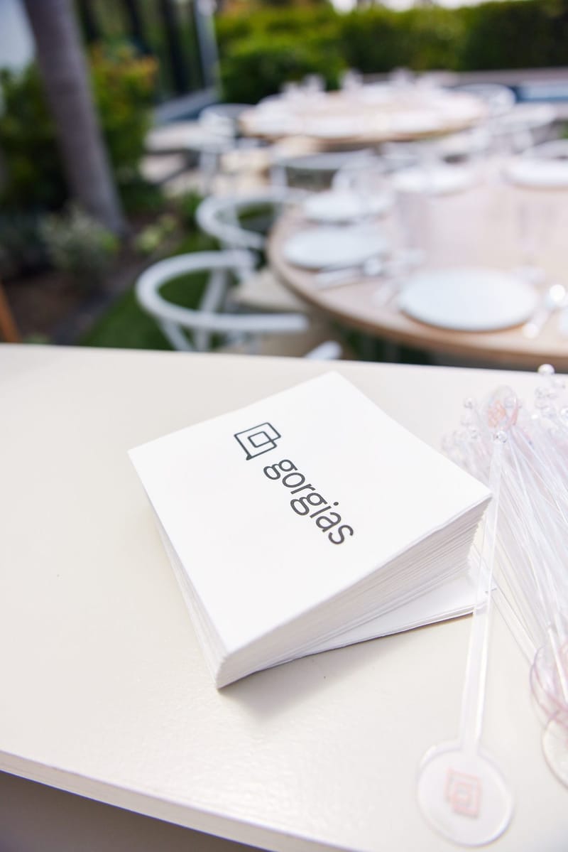 Stack of napkins printed gorgias beside clear stir sticks with the logo, set tables behind