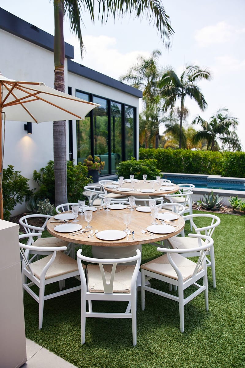 Round table with white wishbone chairs on the lawn by the pool and palm trees