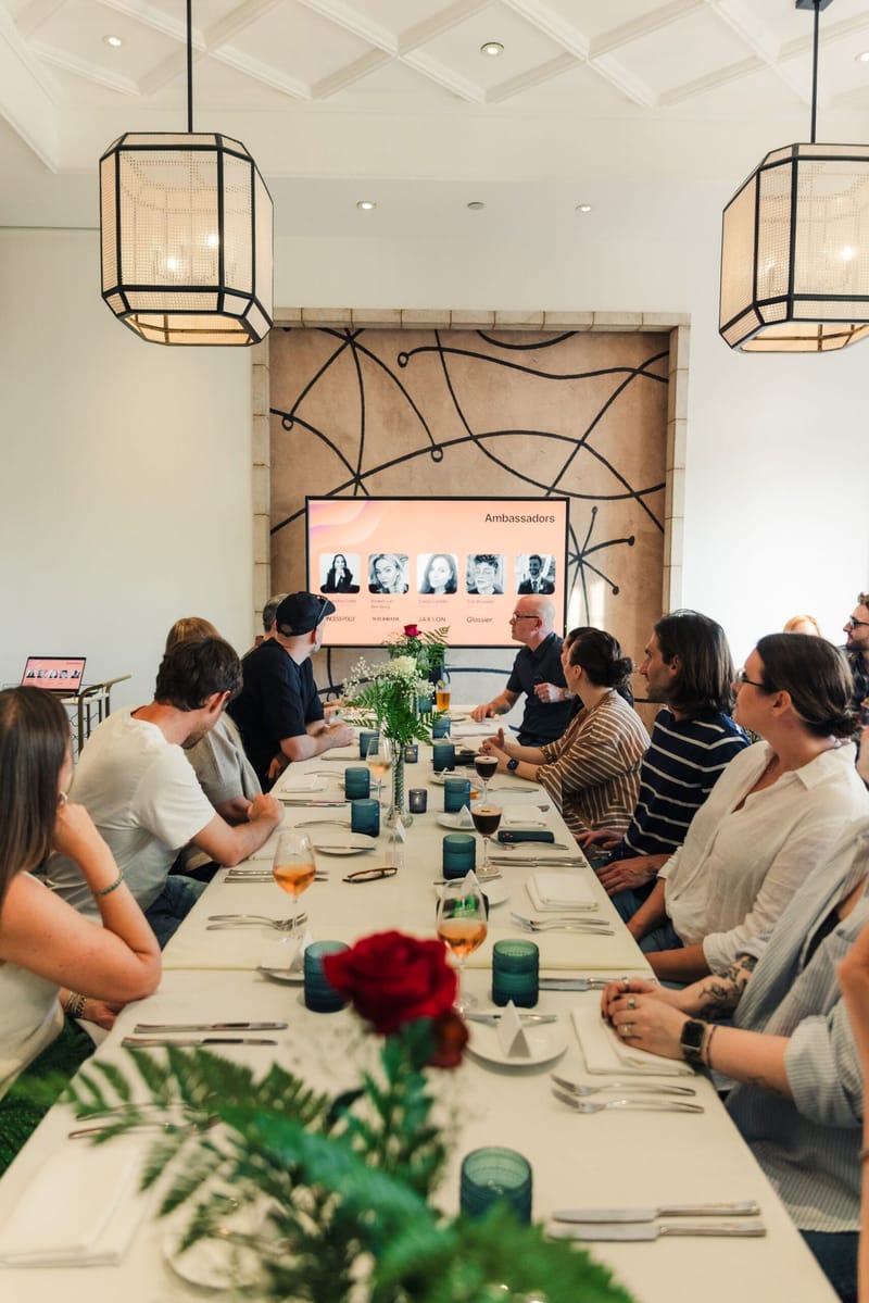 Long dinner table with red roses and teal glassware facing a screen titled Ambassadors with five headshots