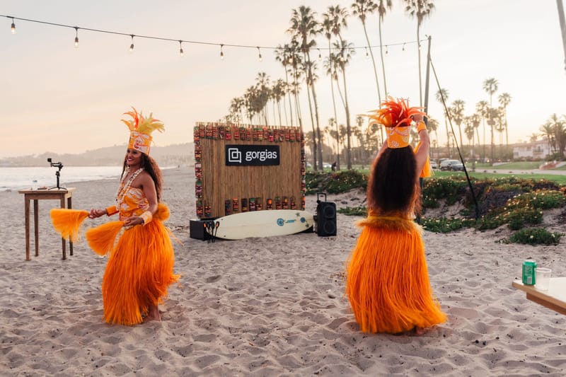 Two dancers in orange grass skirts and feathered headdresses on the beach by the tiki wall and string lights