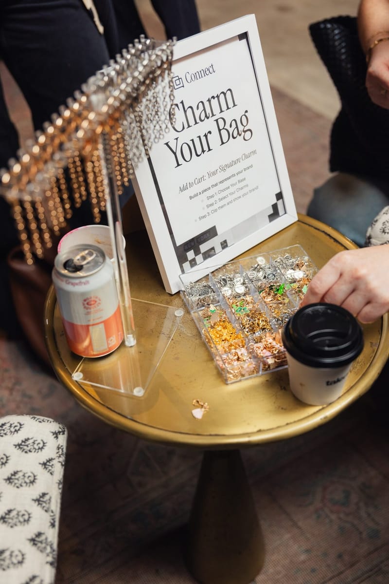 Charm Your Bag sign on a gold tray with bag charms, a drink can and a coffee cup