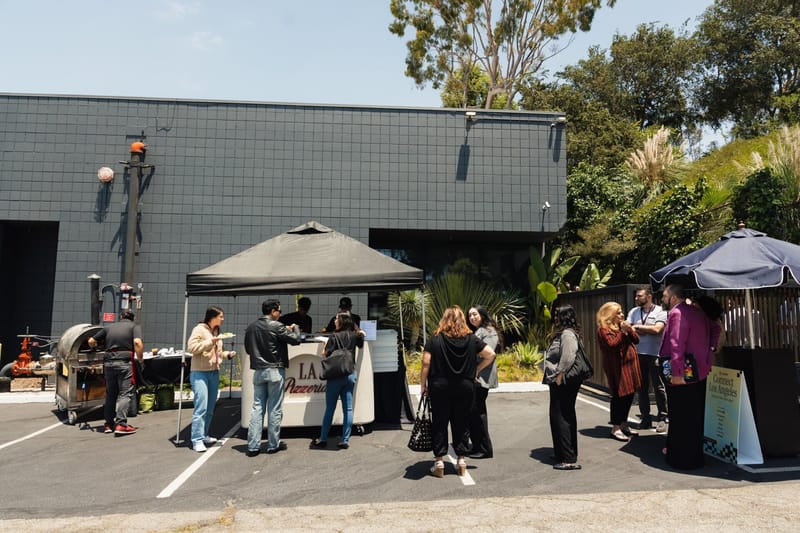 Guests around a white pizza cart lettered Pizzeria under a black pop-up tent in the parking lot, with a wood fired oven on a wheeled trailer beside it